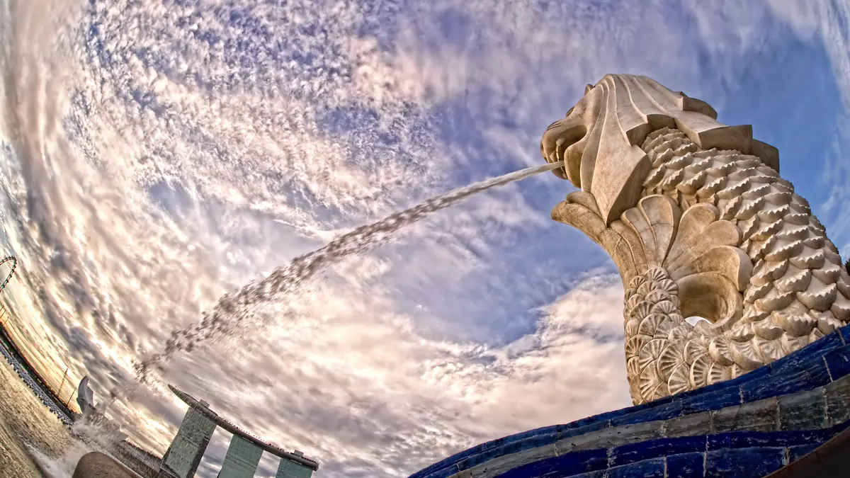 Merlion Park — Looking up at the Merlion statue spouting water against a cloud-streaked sky